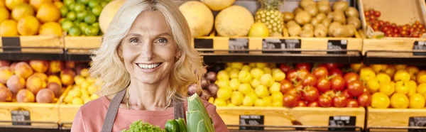 Joyful mature seller with greens smiling at camera with fruits and vegetables on backdrop, banner — Stock Photo