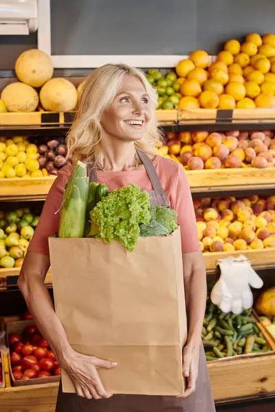 Maturo allegra commessa posa con sacchetto di carta pieno di verdi e guardando lontano a negozio di alimentari — Foto stock