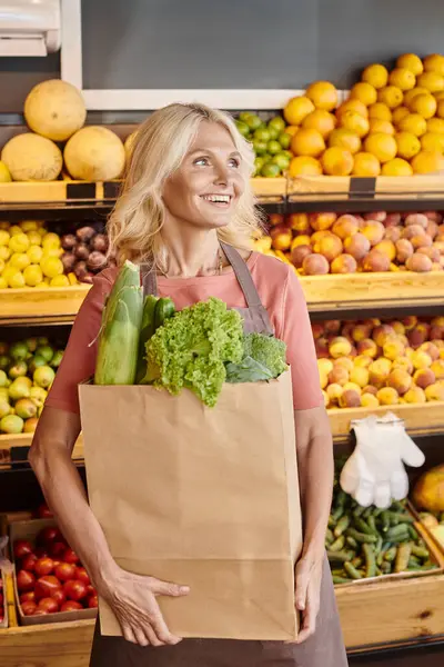 Mature cheerful saleswoman posing with paper bag full of greens and looking away at grocery shop — Stock Photo
