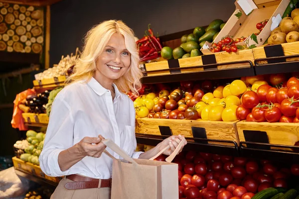 Joyeuse cliente blonde en tenue décontractée posant avec un sac en papier et souriant à la caméra à l'épicerie — Photo de stock