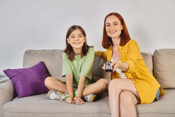 Mulher alegre com controle remoto assistindo tv com filha adolescente no sofá na sala de estar — Fotografia de Stock