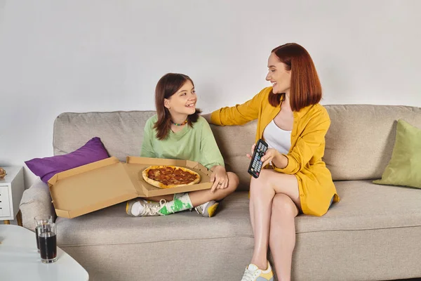 Happy mother and teen daughter watching tv on couch near pizza and glasses with soda in living room — Stock Photo