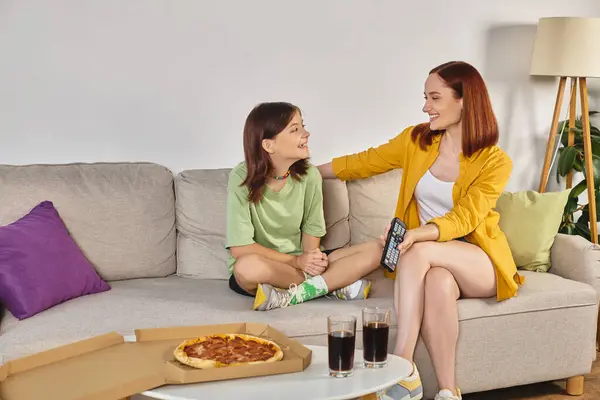 Happy mother and teen daughter watching tv on couch near pizza and glasses with soda in living room — Stock Photo