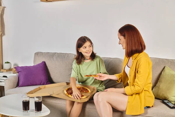 Sonriente mujer dando pedazo de pizza a adolescente hija cerca de gafas con soda en sala de estar - foto de stock