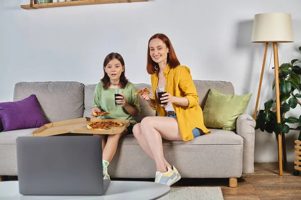 Щаслива жінка з дочкою-підлітком сидить на дивані з піцою і содовою і дивиться фільм на ноутбуці — стокове фото
