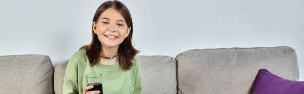 Alegre adolescente sosteniendo vaso de refresco mientras está sentado en el sofá en casa, pancarta - foto de stock