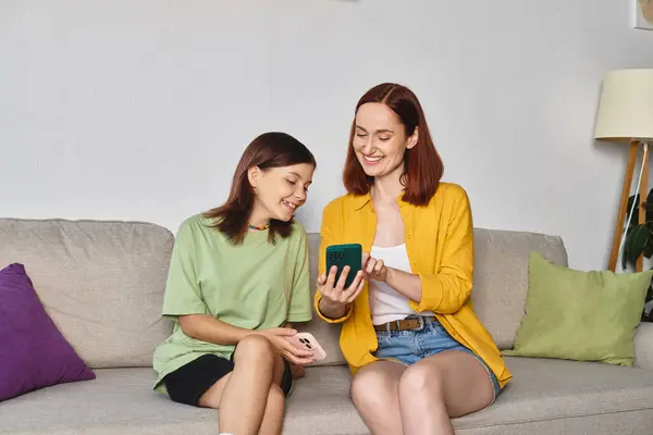 Cheerful woman showing mobile phone to smiling teenage daughter on cozy couch in living room — Stock Photo
