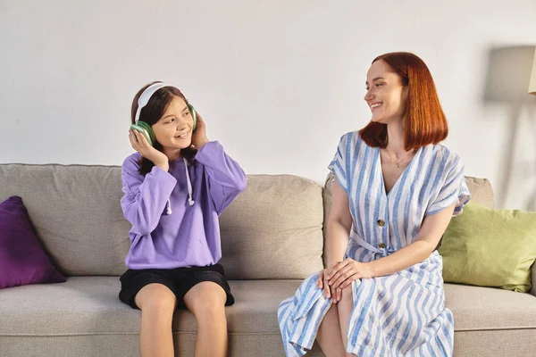 Радісна дівчина-підліток слухає музику в бездротових навушниках біля усміхненої матері на дивані вдома — стокове фото