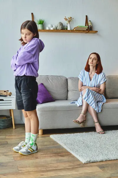 Ображена дівчина-підліток стоїть зі складеними руками біля незадоволеної матері на дивані, сімейний конфлікт — стокове фото