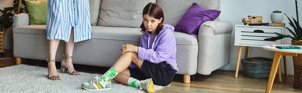 Ображена дівчина-підліток сидить на підлозі у вітальні біля стоячої матері, розрив покоління, банер — стокове фото