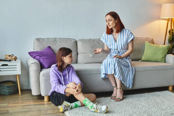 Засмучена жінка розмовляє з ображеною дочкою-підлітком, що сидить на підлозі у вітальні, розрив покоління — стокове фото