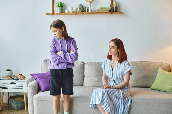 Offended teenage girl standing with folded arms near upset mother sitting on couch, generation gap — Stock Photo