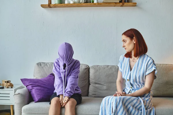 Тривожна жінка, яка розмовляє з засмученою дочкою, затемнює обличчя з капюшоном на дивані у вітальні, конфлікт — стокове фото