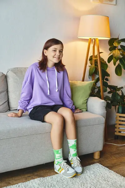 Cheerful teenage girl sitting on cozy couch in living room and smiling at camera, domestic scene — Stock Photo