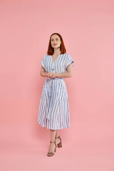 Full length of stylish redhead woman in striped dress standing and looking at camera on pink — Stock Photo