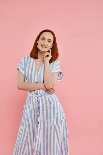 Gaie rousse femme en robe rayée regardant la caméra sur fond rose, portrait studio — Photo de stock