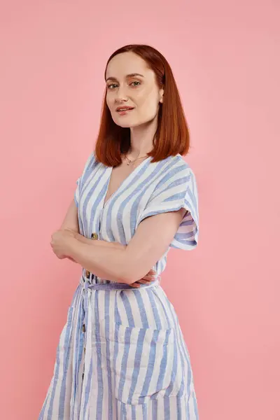 Femme confiante en robe rayée posant avec les bras croisés et regardant la caméra sur fond rose — Photo de stock