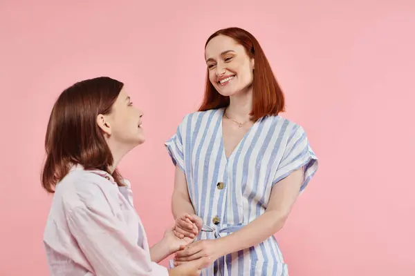 Joyful and stylish woman with teen daughter holding hands and smiling at each other on pink — Stock Photo