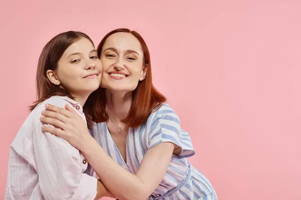 Mujer alegre con hija adolescente en elegante atuendo casual abrazando y mirando a la cámara en rosa - foto de stock