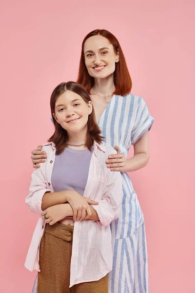 Cheerful and stylish woman hugging shoulders of teenage daughter and looking at camera on pink — Stock Photo