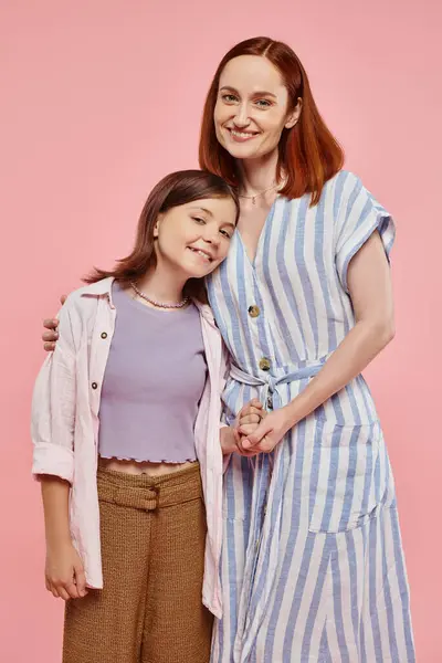 Mujer alegre y elegante con hija adolescente cogida de la mano y sonriendo a la cámara en el telón de fondo rosa - foto de stock