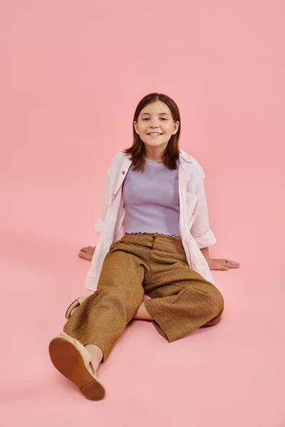 Full length of joyful teenage girl in trendy casual attire sitting and smiling at camera on pink — Stock Photo