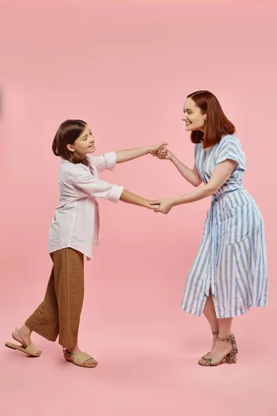 Full length of carefree and trendy woman with preteen daughter holding hands and dancing on pink — Stock Photo