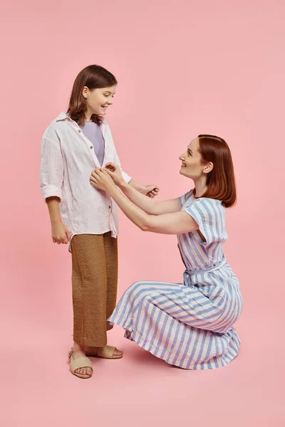Madre sentada en haunches y camisa abotonadora de niña preadolescente sobre fondo rosa, amor y cuidado - foto de stock