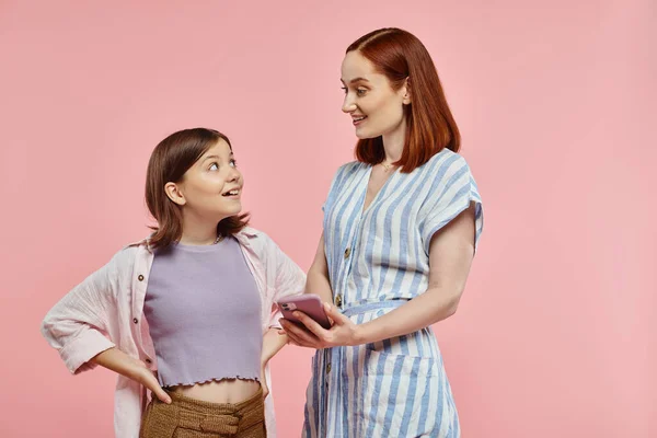 Alegre y elegante madre e hija adolescente mirándose el uno al otro cerca del teléfono inteligente en rosa - foto de stock