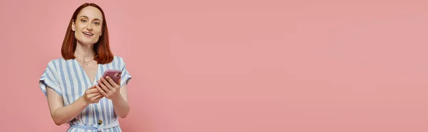 Весела і стильна жінка в смугастій сукні тримає смартфон і дивиться на камеру на рожевому, банер — стокове фото