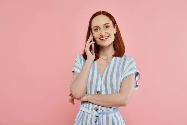 Happy and trendy woman in striped dress talking on smartphone and looking at camera on pink — Stock Photo