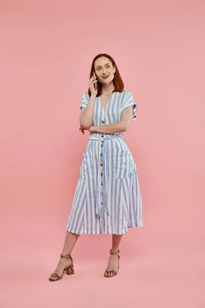 Full length of happy and trendy woman in striped dress talking on smartphone on pink backdrop — Stock Photo