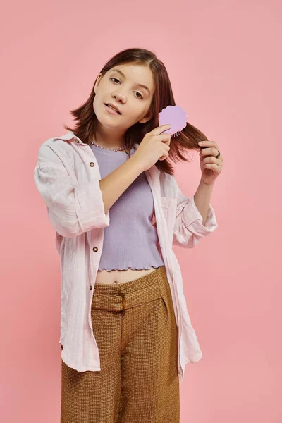 Menina pré-adolescente em roupas elegantes escovando o cabelo e olhando para a câmera no pano de fundo rosa, rotina de beleza — Fotografia de Stock