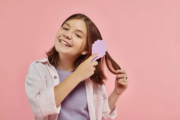 Cheerful preteen girl in stylish clothes brushing hair and looking at camera on pink backdrop — Stock Photo