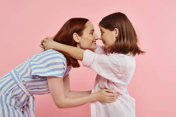 Seitenansicht der fröhlichen Mutter und der schwangeren Tochter, die von Angesicht zu Angesicht auf einer rosa, glücklichen Familie posieren — Stockfoto