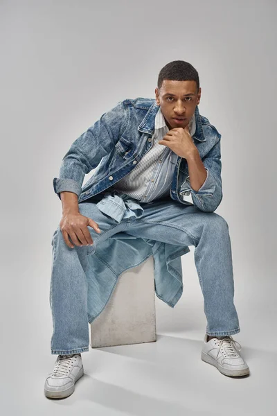 Gut aussehende afrikanisch-amerikanische männliche Modell in Jeans-Outfit sitzt auf White Cube, Modekonzept — Stockfoto