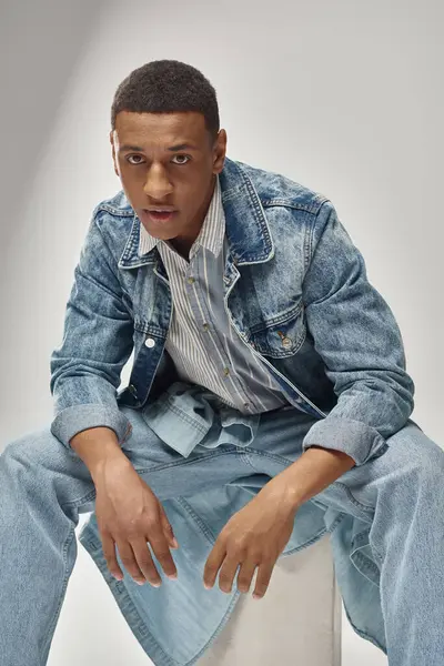 Good looking african american male model in denim outfit sitting on white cube, fashion concept — Stock Photo