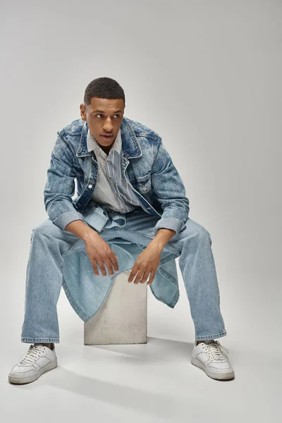 Beau modèle masculin afro-américain en tenue denim assis sur cube blanc, concept de mode — Photo de stock