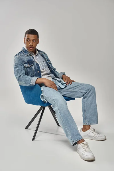 Schöner afrikanisch-amerikanischer Mann in stylischem Jeans-Outfit sitzt auf blauem Stuhl, Modekonzept — Stockfoto