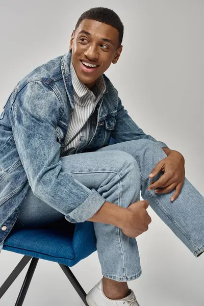 Schöner afrikanisch-amerikanischer Mann in stylischem Jeans-Outfit sitzt auf blauem Stuhl, Modekonzept — Stockfoto