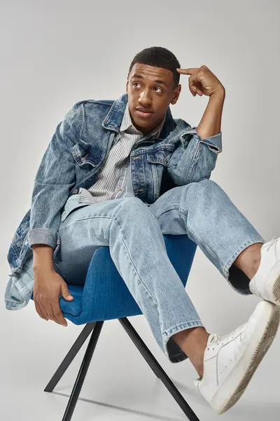 Здивований афроамериканський чоловік у стильному джинсовому вбранні, сидить на синьому стільці, концепція моди — стокове фото