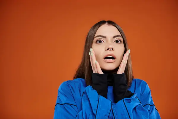 Шокированная женщина с проколотым носом касаясь щек руками и глядя на камеру на оранжевом фоне — стоковое фото