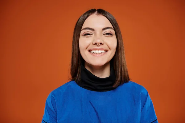 Mujer joven feliz con la nariz perforada mirando a la cámara y sonriendo en el fondo naranja, chaqueta azul - foto de stock