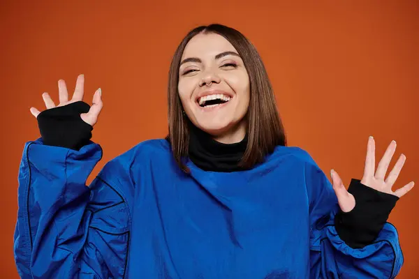 Jeune femme excitée avec nez percé regardant la caméra et souriant sur fond orange, veste bleue — Photo de stock