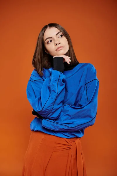 Mujer joven pensativa con la nariz perforada mirando hacia otro lado mientras piensa en fondo naranja, chaqueta azul - foto de stock