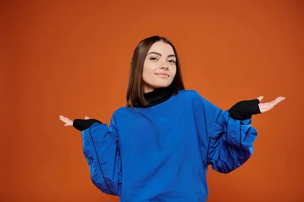 Confused woman with pierced nose looking at camera while standing on orange backdrop, blue jacket — Stock Photo