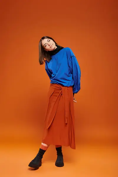 Fall season attire, beautiful woman in skirt and blue sweatshirt standing on orange backdrop — Stock Photo