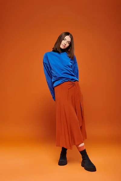 Traje de la temporada de otoño, hermosa mujer en falda y sudadera azul de pie sobre fondo naranja - foto de stock