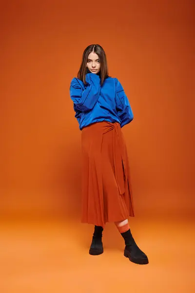 Moda temporada otoño, hermosa mujer en falda y sudadera azul de pie sobre fondo naranja - foto de stock