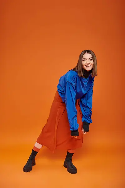 Longitud completa de la joven feliz con la nariz perforada de pie en traje de otoño sobre fondo naranja - foto de stock